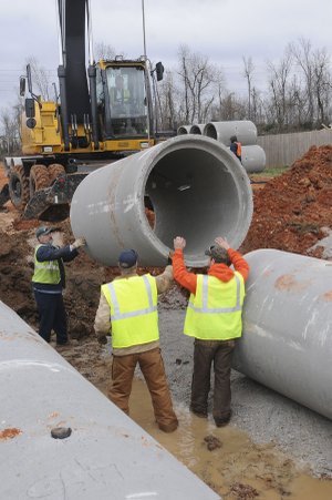 A section of culvert is lowered into place Tuesday by Rogers Street Department superintendent Frankie Guyll, in the track hoe. Jerry Waggoner, from left, Grant Walker and Marc Scott stand by to install it.