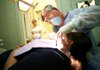Arkansas Democrat-Gazette/RYAN MCGEENEY --07-11-2012-- Angela Heston of Cassville, Mo., 16, left, watches from the hallway as dentist Don Eckard, right, performs a tooth extraction Wednesday on patient Lisa Lynn of Fayetteville in one of the Samaritan Community Center's two cramped dental exam rooms. Heston, a volunteer, has been receiving on-the-job training as a dental assistant through the center. Eckard, who began practicing dentistry in 1972 and is now in self-described "semi-retirement," is one of about a dozen dentists and oral surgeons who volunteer their services for low-income Arkansas residents through the Samaritan Community Center.