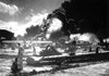 Sailors stand among the wreckage on the airfield at Ford Island Naval Air Station on Dec. 7, 1941, as they watch a ship explode in the background. 